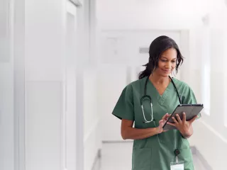 A woman in green scrubs holding a tablet in a hospital hallway