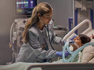 A girl lies in a hospital bed while a healthcare professional attends to her. There is a PrisMax device in the background.