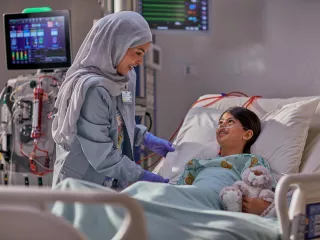 A clinician talks to a young patient who is lying in their hospital bed. There is a PrisMax machine in the background.