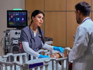 Healthare professional leans over a baby in a bassinet, who is connected to the PrisMax device.